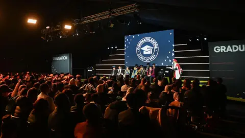 Universidade de sucesso/Greg Brown é uma cerimônia de pós -graduação anterior. Os funcionários e os hóspedes da universidade olham para o estágio em que algumas pessoas se sentam em linhas. Uma pessoa vestindo um boné e um vestido fala em um microfone em pé no palco. 