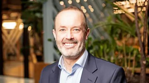 Amazon John Boumphrey, a middle-aged man wearing a suit jacket and shirt. He is smiling, looking at the camera, in front of some plants