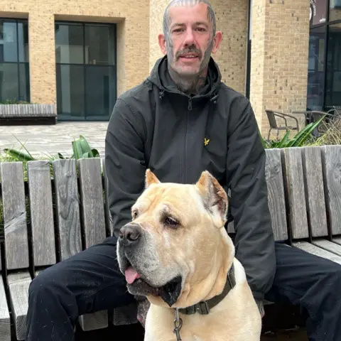 BBC Gareth Thomas is pictured with his dog Swayze. Gareth is sat on a bench wearing black trousers and a black hoodie. He has tattoos on his head and face. Swayze is a Cane Corso with beige fur. 