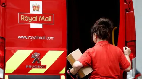 A female postal worker holds a parcel in one arm and closes a door on the back of a red Royal Mail van with the other