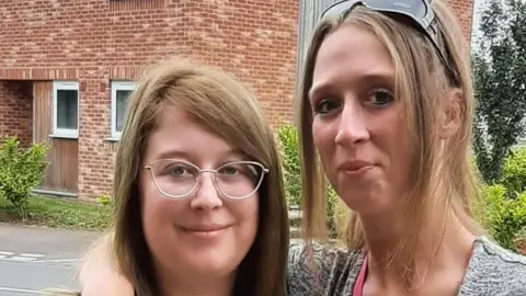 Two women standing outside a brick building. The woman on the right has her arm around the other woman. 
