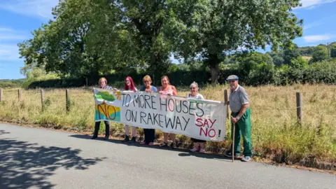 LDRS Six people standing on the edge of a road are holding a large banner that states: "No to more houses on Rakeway. Say no!" There is a field and large trees behind them.