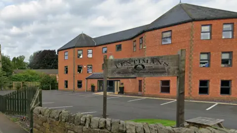 A wooden sign reading FENTIMANS Est 1905 sits behind a short stone fence. There is a three-storey red brick building behind the sign with about a dozen car parking spaces.