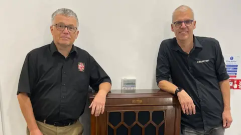 Wayne Bavin/BBC Two men wearing glasses and black shirts are leaning on a wooden piece of furniture.