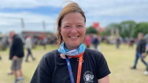 BBC Jodie Rogers has light brown hair tied back. She is wearing a black t-shirt, a blue patternered snood, and a TT lanyard around her neck. She is smiling standing outside with crowds behind her on a sunny and cloudy day.