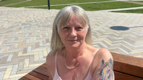 Christine. She has blonde hair with a fringe, wearing a white vest and necklace.