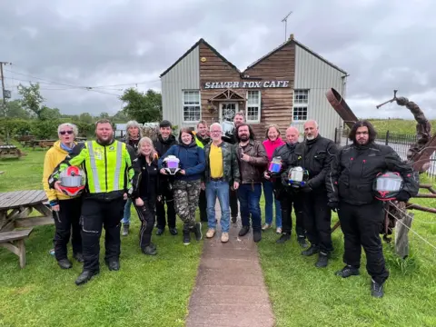 A group of men and women outside a cafe that has the words "silver fox cafe" written on the front in big white letter. Some of the people in the picture are holding motorcycle helmets.