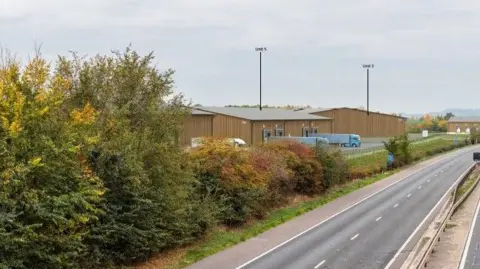 Black Box Planning Partial photo of Brockeridge Farm business centre, obscured by some tress & bushes. On the other side of the bushes is a road.