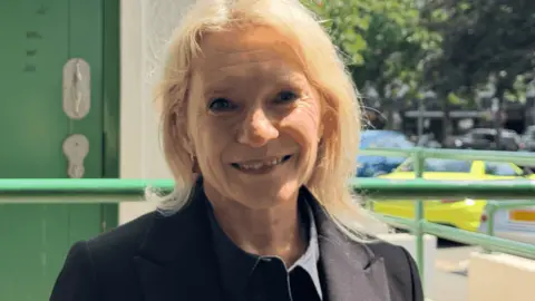 Senator Elaine Millar wearing a dark jacket, standing outside a public toilet entrance, with a green accessible restroom door visible in the background.