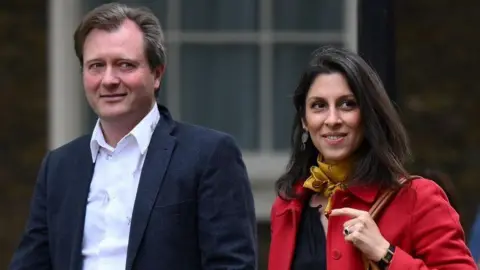 Getty Images Richard Ratcliffe, wearing a white shirt and dark jacket, with Nazanin Zaghari-Ratcliffe, wearing a red coat with a yellow neckerchief, pictured outside as they walk next to each other in Downing Street