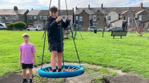 Ethan and Kaelan pictured in the park. One of the boys is standing on a large circular 'bird's nest' swing made from ropes. He is holding onto the swing's chains for balance.