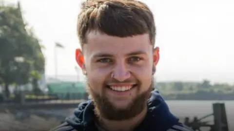 Family photo Connor Marshall smiling at the camera. He has short dark hair, a dark beard and has a nose ring.