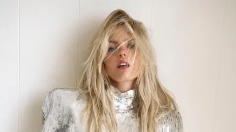 Zora Sicher Promotional photo of Renée Rapp shows her against a plain background, wearing a silver sequinned top, with white jogging pants. Her hair is artfully messed up, and she holds her left hand across an exposed midriff