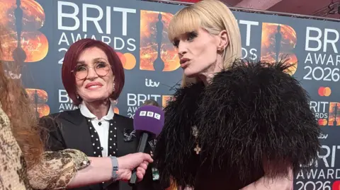 BBC A woman with red hair and a woman with blonde hair and a fur coat, stand on a red carpet speaking into a microphone.