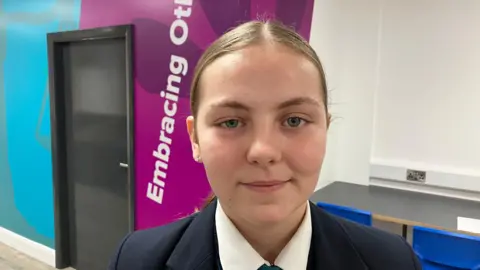 A school girl, pictured from the shoulders up. She is wearing a shirt and a blazer. She has blonde hair, tied back. She is standing inside the school, with a white, purple and blue wall behind her. 