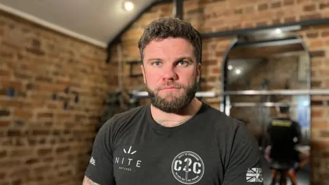 Head and shoulders shot of David Coleman sat down in a small gym with bare brick walls, he looks directly at the camera and is wearing a dark grey t-shirt.