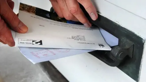 A hand putting letters into a black letterbox that is close to ground level.

