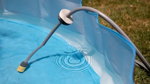 Getty Images A hosepipe is laying in a blue paddling pool which is sitting on grass. Water has filled up the pool half way.