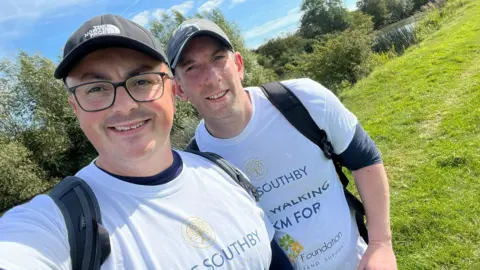 Dave Southby Dave Southby and his colleague Neil Sawyer posing for a selfie beside a river in a grassy field. They are both wearing caps and white t-shirts which say 'Dave Southby Financial Planning. We're walking 100km for the Kelly Foundation.'