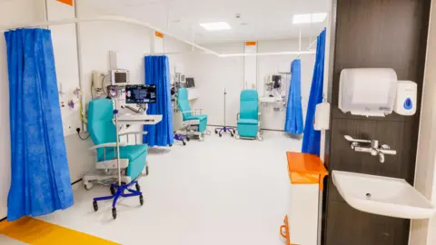 PHU A clean-looking medical room with blue paper curtains and three blue recliner chairs in it. In the foreground, outside the ward, is a sink attached to the wall with soap and paper towels.