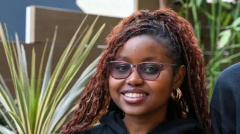 ISCRE A young woman smiles for the camera. She is standing in front of a large plant. She is wearing her hair in a ponytail and has glasses on and gold hoop earrings. 