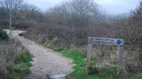 N Chadwick A sign beside a path curving through bare shrubs and trees in winter reads "Bothenhampton Nature Reserve"