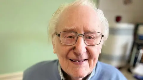 A headshot of Eric Page who is wearing square-framed eye glasses and a blue jumper.