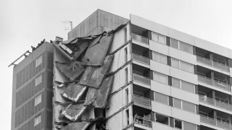 PA Media A black and white image showing several collapsed floors on the corner of the Ronan Point building. The two sides, floors and ceilings have fallen, with some concrete panels still hanging off the remaining structure