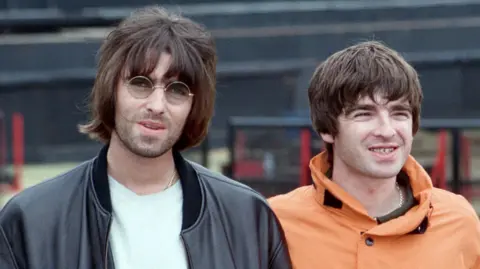 Getty Images Liam and Noel Gallagher as seen in the '90s. Liam has long black hair, stubble and round framed glasses and is wearing a black leather jacket. Noel is smiling and has short black hair and is wearing an orange parka coat.