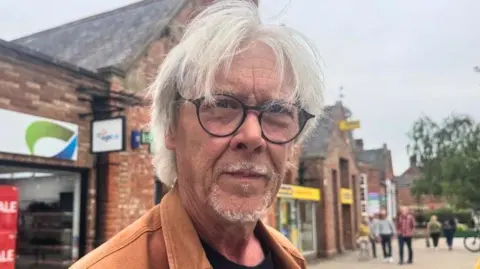 BBC/ Emily Johnson A man with grey hair and glasses stands on a high street in Acomb, York.