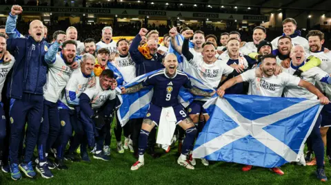 SNS The Scotland men's football team, posing for a victory photo on the pitch at Hampden Park. The men are cheering, their arms aloft, with a jubilant John McGinn in the front row holding a saltire flag.