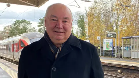 Dennis Fancett is in his 60s wearing a blue jacket with a scarf tucked in. He is standing on a railway platform and behind you can see an LNER train approaching 