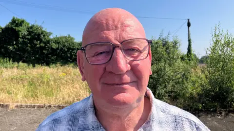 Terry Crossan has black-rimmed glasses, and is wearing a blue and black chequered shirt. He is standing near a grassy area.