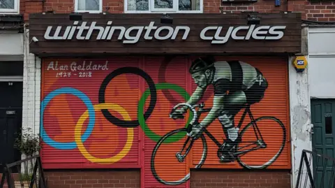 Withington Walls Alan Geldard on his bike aginst the Olympic rings painted on a shop shutter in Withington