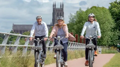 The council’s deputy director for policy and strategy David Sutton and corporate director for planning and governance Sian Stroud with Beryl CEO Phil Ellis. The trio are riding bikes along the river with a metal barrier to their right and Worcester Cathedral in the backdrop.