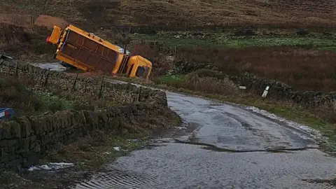 Andrew Hall An overturned gritting lorry stuck in a ditch off a road on great, icy, wet moorland countryside