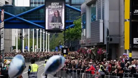 PA Media Crowds of people behind metal barriers at the side of a main road leaning over. The road is clear, but a sign above the road features an image of Ozzy Osbourne, with Birmingham will always love you written underneath.