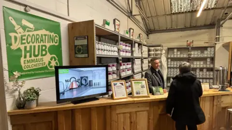 A large warehouse has racks of shelving covered in paint tins and other supplies. A large green sign on the wall says it is a Decorating Hub. Two people wearing winter coats stand and talk to each other.