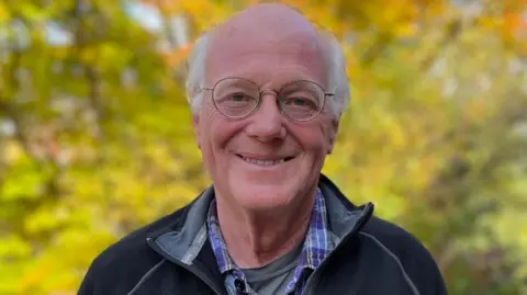 Ben Cohen Ben has receding grey hair and is wearing metal-rimmed specs on his face, a black jacket over a checked shirt. He is standing in a forest.