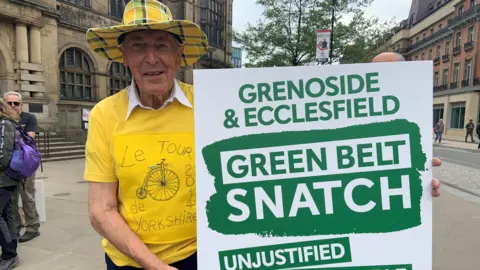 An elderly man wearing a yellow check hat and a yellow t-shirt with the words Le Tour de Yorkshire written on it. He is holding a green-and-white placard.