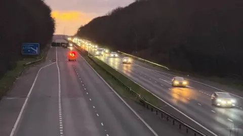 A motorway with vehicles travelling towards the camera on the right. There is no traffic on the left carriageway. The road is lined with trees and the sun is going down in the background