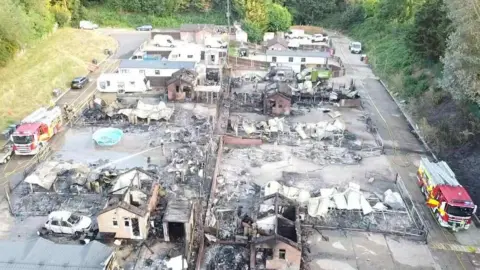 Three Counties Guttering & Roofing An aerial view of a large fire-damaged area with multiple destroyed camping pitches and vehicles. There is extensive damage with charred remains of structures and debris scattered around. Two fire engines are on the site, one on the left side and one on the right.