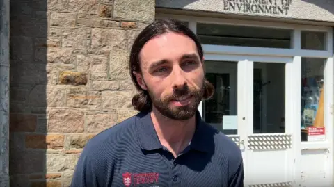 Alex Plaster has long dark hair pulled back and a short beard, wearing a navy polo shirt, standing outdoors in front of a stone building. A white-framed glass door is behind him, and part of a sign above the entrance reads “ENVIRONMENT.” The scene is lit by bright sunlight, casting strong shadows on the wall.