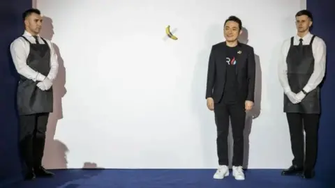 Getty Images A young man poses next to a large white board, which has a banana taped to it with duct tape. Two auction handlers wearing black aprons over a white shirt flank the board. The man, Justin Sun, wears black trousers with a black t shirt and black jacket with white trainers and is smiling