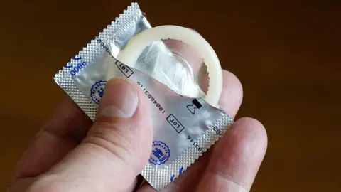 A hand is holding an open silver foil condom wrapper, with a condom exposed.