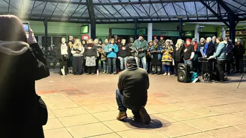 Maddy Jennings/BBC About 30 people standing in a line singing. A man rests on one knee as he watches the performance while a woman films it on her phone