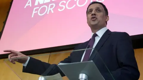 PA Media Anas Sarwar with dark hair, wearing a dark suit, white shirt and purple tie, points to his right while speaking on stage. He is photographed from a low angle, standing behind a podium and in front of a red Labour sign. 