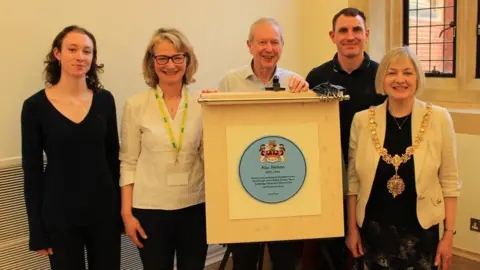 Cambridge Past, Present & Future Five people are standing next to each other in the corner of a room. A man, who is in the middle, is holding a wooden board which has a blue plaque on it. 