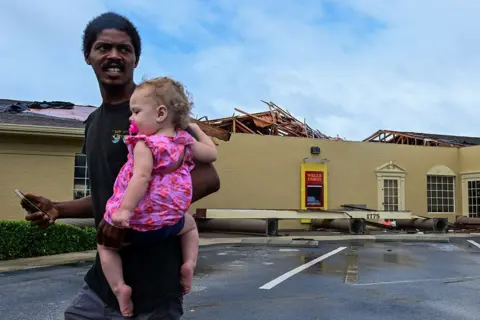 Giorgio Viera / AFP A man walks while carrying a baby with a Wells Fargo bank branch in the background with its roof in splinters