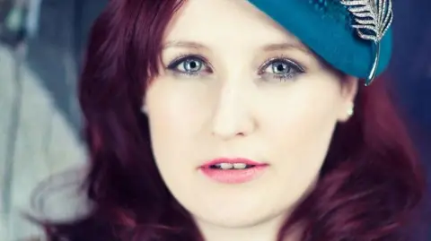 Lou Hickey looks into the camera - she is a woman with dark hair and a turquoise hat on 